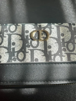 Plånbok cd - Snygg plånbok från CD med klassiskt monogrammönster i svart och grått tyg, detaljer i svart läder och guldfärgad CD-logga framtill. Plånboken har guld dragkedja och stilren design, perfekt för att hålla koll på kort och cash.