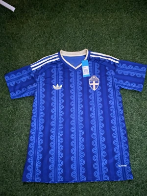 Blå Sverige fotbollströja Adidas - Säljer en blå fotbollströja från Adidas med Sveriges landslagsemblem på bröstet och vita detaljer. Tröjan har ett unikt mönster i olika blå nyanser, vita ränder på axlarna och V-ringad krage. Materialet är lätt och andas, perfekt för träning eller match.