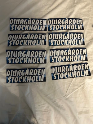 Djurgården Stockholm klistermärken - Säljer åtta stycken klistermärken med texten 'Djurgården Stockholm' i vit och blå färg på svart bakgrund. Perfekta för att visa sitt stöd för Djurgården, kan fästas på olika ytor. Rektangulär form och tydlig text.