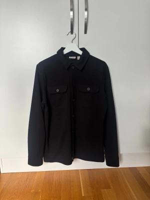 Svart overshirt från Only and Sons - Stilren svart overshirt från Only and Sons med klassisk krage, knäppning framtill och två bröstfickor med lock och knapp. Långärmad modell i ett mjukt och lite tjockare tyg som passar perfekt som lager på lager.