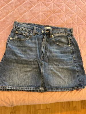 Blå jeanskjol med fem fickor - Klassisk blå jeanskjol i kort modell med fem fickor och bälteshällor. Kjolen har dragkedja och knapp framtill och är tillverkad i denim. Perfekt för en avslappnad och trendig look.