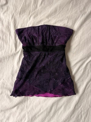 Lila spets topp med svart knytband - En kort lila topp med dekorativ spets och svart knytband i midjan. Toppen är axelbandslös och har ett vackert mönster i spetsen. Perfekt för dig som vill ha en unik och färgstark look. Materialet känns lätt och bekvämt mot huden.