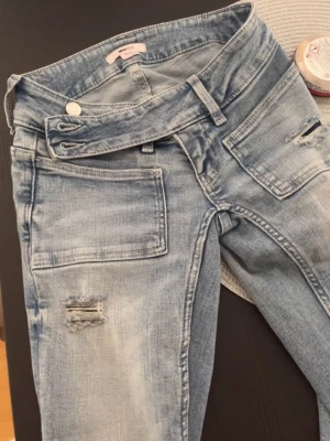 Lågmidjade bootcut jeans  - Säljer ett par ljusblå bootcut jeans med coola slitningar vid knäna och råa detaljer. Jeansen har hög midja, breda framfickor och en unik asymmetrisk knäppning. Perfekta för dig som gillar en trendig och avslappnad stil. Storlek 14 + XS lite korta på mig som är 170