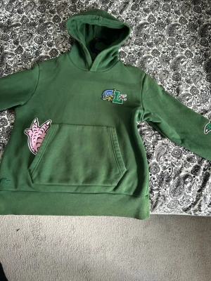 Lacoste hoodie  - Säljer en grön hoodie med coola tryck på bröstet, magen och ena ärmen. Hoodien har en stor känguruficka framtill och en klassisk huva. Perfekt för dig som gillar streetwear och vill sticka ut lite extra.