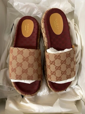 Gucci beige tofflor med GG-mönster - Snygga beige tofflor från Gucci med klassiskt GG-monogram i brunt över hela ovandelen och sidorna. Innersulan är vinröd med diskret GG-mönster och guldfärgad logga. Plattformssula i gummi ger extra höjd och cool look. Perfekta för dig som gillar lyxiga detaljer.