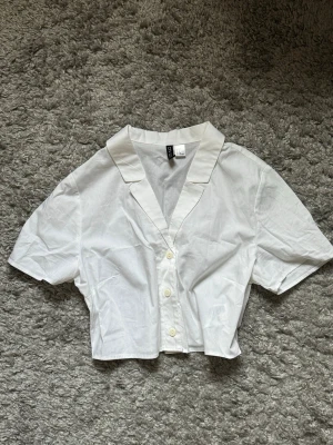 Vit kortärmad blus från H&M - Stilren vit blus från H&M med korta ärmar och klassisk krage. Blusen är croppad och har knäppning framtill med fyra vita knappar. Tillverkad i ett lätt och tunt bomullstyg som känns fräscht och luftigt.