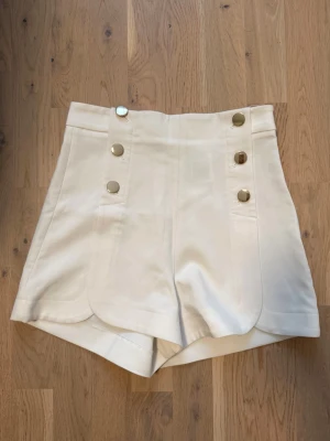 Vita högmidjade shorts med knappar - Snygga vita högmidjade shorts med dekorativa guldknappar framtill. Shortsen har en unik skuren kant nedtill och är tillverkade i ett mjukt tyg som känns lyxigt. Perfekta för sommarens alla tillfällen och ger en chic look.
