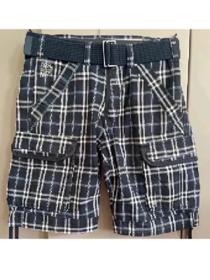 Rutig cargo shorts med bälte - Snygga mörkblå och vita rutiga cargo shorts från JICRe med praktiska fickor och tillhörande tygbälte. Perfekta för sommaren med avslappnad passform och coolt mönster. Broderad logga på ena fickan ger extra detalj.