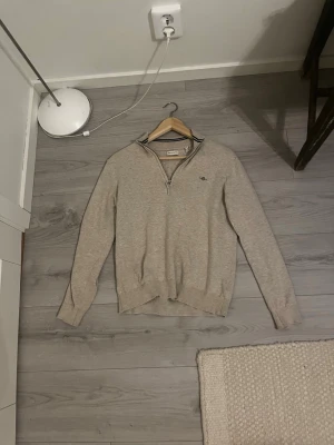 Beige halvzip-tröja från GANT - Snygg beige stickad tröja från GANT med halv dragkedja och liten broderad logga på bröstet. Tröjan har ribbade muddar och krage med mörkblå ränder på insidan. Perfekt för en stilren och avslappnad look.