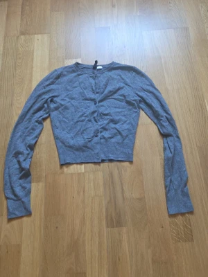 Grå cropped kofta med knappar - Grå, tunnstickad cropped kofta med långa ärmar och rund halsringning. Koftan har knäppning framtill med små knappar och är i ett mjukt, stretchigt material. Perfekt att slänga över en topp eller klänning.