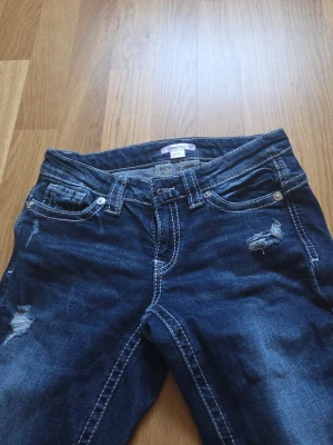 Mörkblå bootcut jeans med slitningar - Snygga mörkblå bootcut jeans från Gina Tricot med slitna detaljer på låren och knäna. Jeansen har klassisk femficksmodell, vita kontrastsömmar och låg midja. Perfekta för en avslappnad och trendig look.