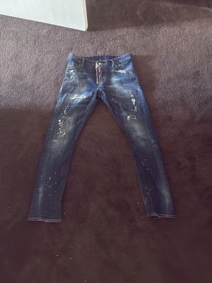 Mörkblå Dsquared2 jeans med zipdetalj - Säljer ett par mörkblå jeans från Dsquared2 med cool zipdetalj och slitningar. Jeansen har skinny fit, målade stänk och slitna partier för en trendig, avslappnad look. Perfekta för dig som gillar streetstyle och vill sticka ut.