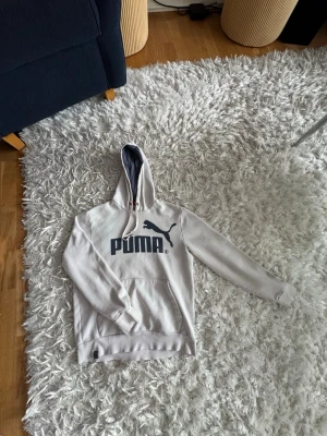 Vit hoodie från Puma med logga - Snygg vit hoodie från Puma med stor svart logga och text på bröstet. Hoodien har en klassisk känguruficka framtill och justerbar huva med grått innerfoder. Perfekt för en avslappnad och sportig stil.