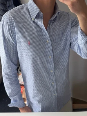 Blårutig skjorta från Polo Ralph Lauren - Klassisk blårutig skjorta från Polo Ralph Lauren med button-down krage och broderad logga på bröstet. Skjortan har långa ärmar, vita knappar och är tillverkad i mjuk bomull. Perfekt för en stilren look. Storlek 39 vilket motsvarar m. Den är lite hålig i kragen som man ser på sista bilden, därav priset. Annars bra skick