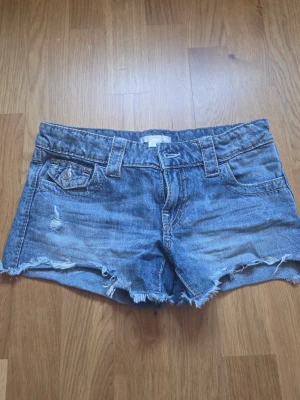 Blå jeansshorts med fransig kant - Snygga blå jeansshorts med fransig kant och slitna detaljer. Klassisk femficksmodell med bälteshällor och knappstängning. Perfekta för varma dagar och en avslappnad stil.