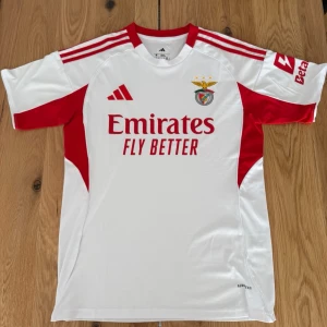 Benfica matchtröja Adidas vit/röd - Snygg Benfica matchtröja från Adidas i vitt med röda detaljer och logga. Tröjan har klubbmärke, sponsortryck och klassiska Adidas-ränder på axlarna. Perfekt för fotbollsträning eller att bära på läktaren.
