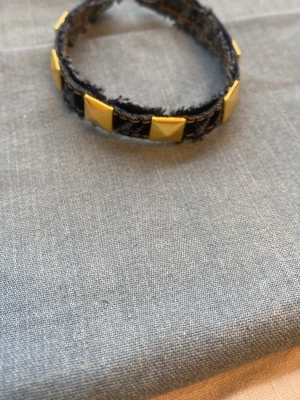 Mörkblått denimarmband med guldnitar - Coolt armband i blått denimtyg med råa kanter och fyrkantiga guldiga nitar runt om. Perfekt accessoar för att ge din outfit en edgy touch. Armbandet har en trendig och ungdomlig design som sticker ut.