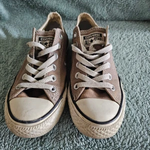 Beige Converse All Star låga sneakers - Säljer ett par klassiska Converse All Star sneakers i beige canvas med vita gummisulor och tåhätta. Skorna har vita snören, metallöljetter och den ikoniska loggan på plösen. Perfekta för en avslappnad och stilren look.