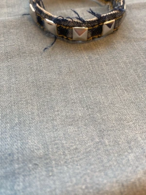 Jeansarmband med nitar - Snyggt armband i mörkblå denim med fransiga kanter och silvriga fyrkantiga nitar runt om. Perfekt accessoar för en cool och avslappnad stil. Justerbar storlek med tryckknapp.