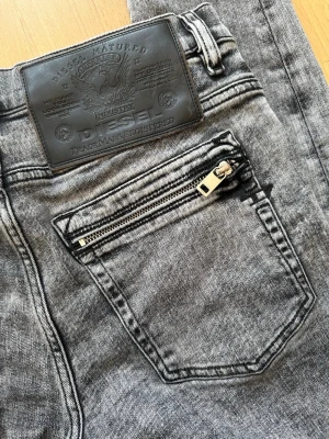 Diesel jeans - Gråa slimjeans från Diesel med tvättad look och dragkedjedetalj på bakfickan. Klassisk femficksmodell med svart läderpatch bak i midjan. Storlek 32💫