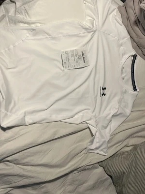 Vit Under Armour t-shirt - Säljer en vit t-shirt från Under Armour med svart logga på bröstet. Tröjan är tillverkad i ett tekniskt material som är perfekt för träning och sport. Den har korta ärmar och en rund halsringning. Perfekt för dig som vill ha en snygg och funktionell sporttröja. Helt ny endast provad en gång 