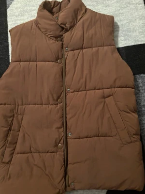 Brun dunväst från H&M - Snygg brun dunväst från H&M med hög krage och tryckknappar framtill. Västen har två stora fickor på sidorna och är quiltad för extra värme. Perfekt att slänga över en hoodie när det är kyligt ute. Snabb leverans, storleken finns i en av bilderna. 