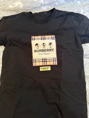Svart Burberry t-shirt med rutigt tryck - Snygg svart t-shirt från Burberry med klassiskt rutigt tryck och logga på bröstet. T-shirten har rund hals och korta ärmar. Perfekt för dig som gillar exklusiva märken och stilren design.