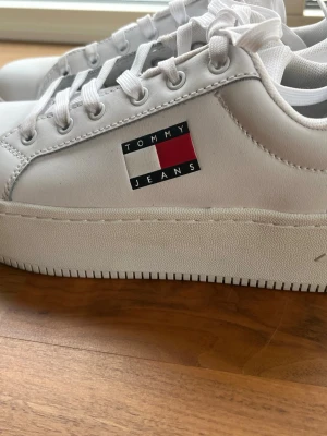 Vita sneakers från Tommy Jeans - Fräscha vita sneakers från Tommy Jeans med klassisk logga i rött, vitt och blått på sidan. Skorna har lågt skaft, vita snören och en chunky sula. Tillverkade i slätt syntetmaterial med textilfoder för extra komfort.
