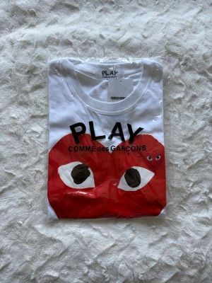 CDG vit hjärta t shirt  - Vit t-shirt från Comme des Garçons Play med det ikoniska röda hjärtat med ögon tryckt på bröstet. Klassisk passform och rund halsringning. Perfekt för dig som gillar streetstyle och vill sticka ut med en cool grafisk detalj. Passar kanske lite mer åt S sidan. Har aldrig använts 