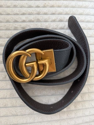 Svart skärp med GG-spänne från Gucci - Snyggt svart skärp från Gucci med det ikoniska dubbla GG-spännet i guld. Bältet är gjort i skinn och har en klassisk, stilren design som passar till det mesta. Perfekt accessoar för att lyfta vilken outfit som helst.