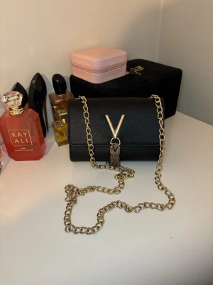 Svart axelväska från Mario Valentino - Snygg svart axelväska från Mario Valentino med guldfärgad kedja och V-detalj framtill. Perfekt storlek för det viktigaste och ger en lyxig touch till din outfit. Hör gärna av er vid frågor och funderingar😊🫶🏼