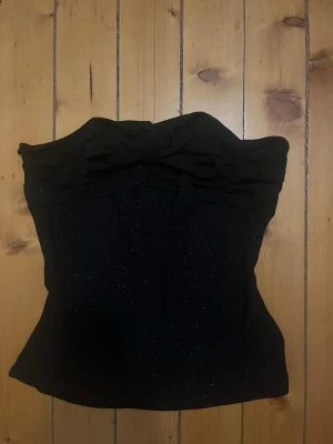 Svart glittrig tubtopp med knyt - Snygg svart tubtopp med glittriga detaljer och knytband i nacken. Toppen har en söt rosettdetalj framtill och är figurnära i modellen. Perfekt för dig som vill sticka ut lite extra.
