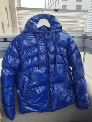 Blå pufferjacka från Guess, storlek XS - Snygg blå pufferjacka från Guess med glansig finish och huva. Jackan har dragkedja framtill, två stora fickor och justerbara snören vid huvan med Guess-logga. Perfekt för kalla vinterdagar och ger en cool streetstil. Storlek XS. Har inte använts, helt ny.