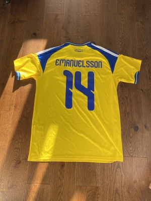 Svensk landslagströja Adidas gul/blå - Gul svensk landslagströja från Adidas med blå detaljer, nummer 14 och namnet Emanuelsson på ryggen. Tröjan har korta ärmar, blå ränder på axlarna och det svenska landslagsemblemet på bröstet. Perfekt för fotbollsfans!
