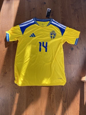 Svensk fotbollströja Adidas gul/blå - Säljer en gul och blå fotbollströja från Adidas med Sveriges landslagsemblem och nummer 14 på både fram- och baksidan. Tröjan har klassiska Adidas-ränder på axlarna och är tillverkad i ett lätt och svalt polyestermaterial. Perfekt för match eller träning!