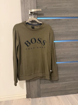 Olivgrön sweatshirt från Hugo Boss - Cool olivgrön sweatshirt från Hugo Boss med stort BOSS-tryck på bröstet. Tröjan har rund halsringning, långa ärmar och ribbade muddar. Materialet känns mjukt och bekvämt, perfekt för en avslappnad look.