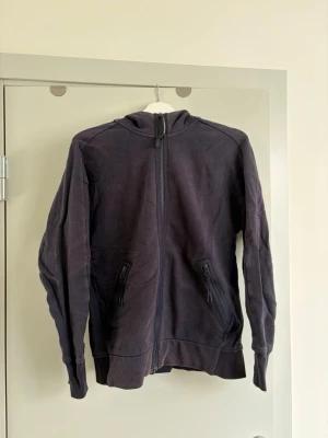 Mörkblå hoodie med glasögon i huvan - Säljer en mörkblå hoodie från C.P. Company med dragkedja framtill och två fickor med dragkedja. Huvan har inbyggda glasögon som ger en unik och cool look. Tillverkad i mjukt bomullsmaterial och har ribbade muddar vid ärmslut och nederkant.