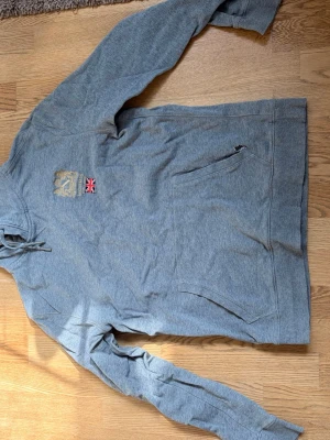 Grå hoodie med brittisk flagga - Grå hoodie med huva och känguruficka framtill. På bröstet finns ett broderat märke i guld och en liten brittisk flagga. Hoodien har dragsko i huvan och är långärmad, perfekt för en chill och avslappnad stil. Finns ett litet hål vid främre fickan och ett litet där bak!