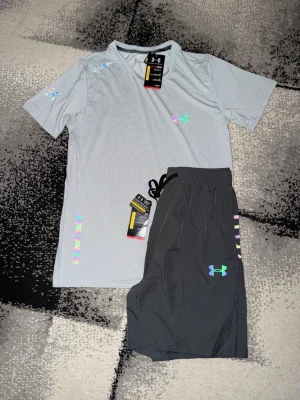 Under Armour träningsset grå/svart - Snyggt träningsset från Under Armour med en ljusgrå t-shirt och svarta shorts. T-shirten har färgglada logodetaljer och korta ärmar. Shortsen har dragsko i midjan och matchande logga. Perfekt för gymmet eller löprundan.