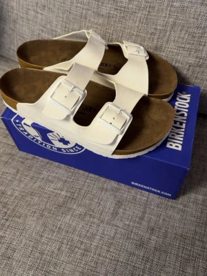 Vita Birkenstock Toflor - Snygga vita sandaler från Birkenstock med två breda remmar och justerbara metallspännen. Fotbädden är i brunt mockaliknande material för extra komfort. Perfekta för en avslappnad och trendig stil.