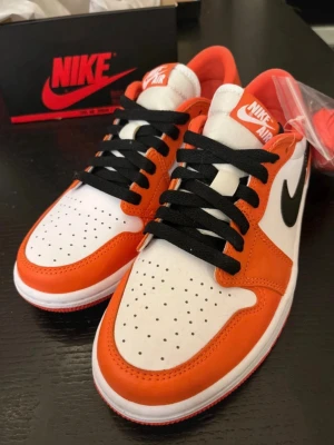Nike Air Jordan 1 Low Orange/White - Säljer ett par Nike Air Jordan 1 Low sneakers i vitt och orange med svarta detaljer. Skorna har klassisk låg siluett, svart swoosh och svarta skosnören. Ovandelen är i läder och tungan har Nike Air-logga. Perfekta för dig som gillar streetwear och snygg design. Box och extra skosnören medföljer!!