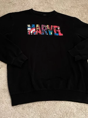 Svart Marvel sweatshirt - Cool svart sweatshirt med färgglad Marvel-logga på bröstet där flera superhjältar syns i bokstäverna. Tröjan har rund hals, långa ärmar och ribbade muddar. Perfekt för dig som älskar Marvel och vill ha en bekväm och snygg tröja till vardags.