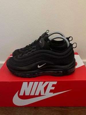 Nike Air Max 97 Triple Black - Nike Air Max 97 Triple Black med ikoniska vågformade paneler, synlig Air Max-sula och små vita Swoosh-loggor på sidan och tungan. Hela skon är svart med detaljer i syntet och mesh för en sportig och clean look.