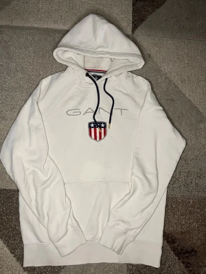 Gant hoodie - M men passar både S och M. Vit hoodie från Gant med broderad logga och amerikanskt inspirerat märke på bröstet. Mörkblå dragsnören med gulddetaljer och klassisk känguruficka. Perfekt för en avslappnad och stilren look. En synligt teat på fikan