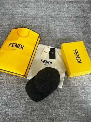 Svart Fendi keps - Säljer en svart keps från Fendi med ikoniskt FF-logomönster över hela kepsen. Klassisk form med böjd skärm och justerbar baksida. Materialet är troligtvis bomull eller canvas, perfekt för dig som vill ha en exklusiv accessoar. Kommer med box och allt. 