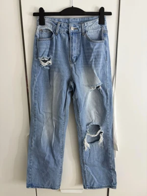 Ljusblå ripped jeans med raka ben - Säljer ett par ljusblå jeans med raka ben och flera slitningar på framsidan. Jeansen har klassisk femficksmodell, hög midja och dragkedja med knapp. Perfekta för en avslappnad och trendig look.