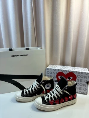 Converse x Comme des Garçons Play High - Säljer ett par Converse x Comme des Garçons Play high-top sneakers i svart canvas med vita detaljer och flera röda hjärtan med ögon på sidan. Klassisk vit gummisula och vita snören. Perfekt för dig som vill sticka ut med en ikonisk och trendig look.