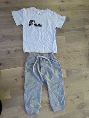 Set med byxa och t shirt  love my mama  - Set med en vit kortärmad t-shirt med texten "LOVE MY MAMA" tryckt på framsidan och ett par grå mjukisbyxor med dragsko i midjan. Byxorna har muddar vid bensluten. T-shirt och mjukisbyxor set "LOVE MY MAMA"