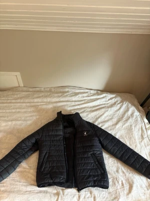 Svart pufferjacka från polo club - Snygg svart pufferjacka från polo club med quiltad design och dragkedja framtill. Jackan har två snedställda fickor med dragkedja och en liten logga på bröstet. Perfekt för kalla dagar och enkel att matcha med olika outfits.