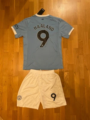 Manchester City Haaland matchställ - Snyggt Manchester City matchställ med Haaland 9-tryck. Tröjan är ljusblå med vita detaljer, klubbmärke och Etihad Airways-logga framtill. Korta ärmar och rund hals. Matchande vita shorts med klubbmärke och nummer 9. Perfekt för fotbollsträning eller fans.